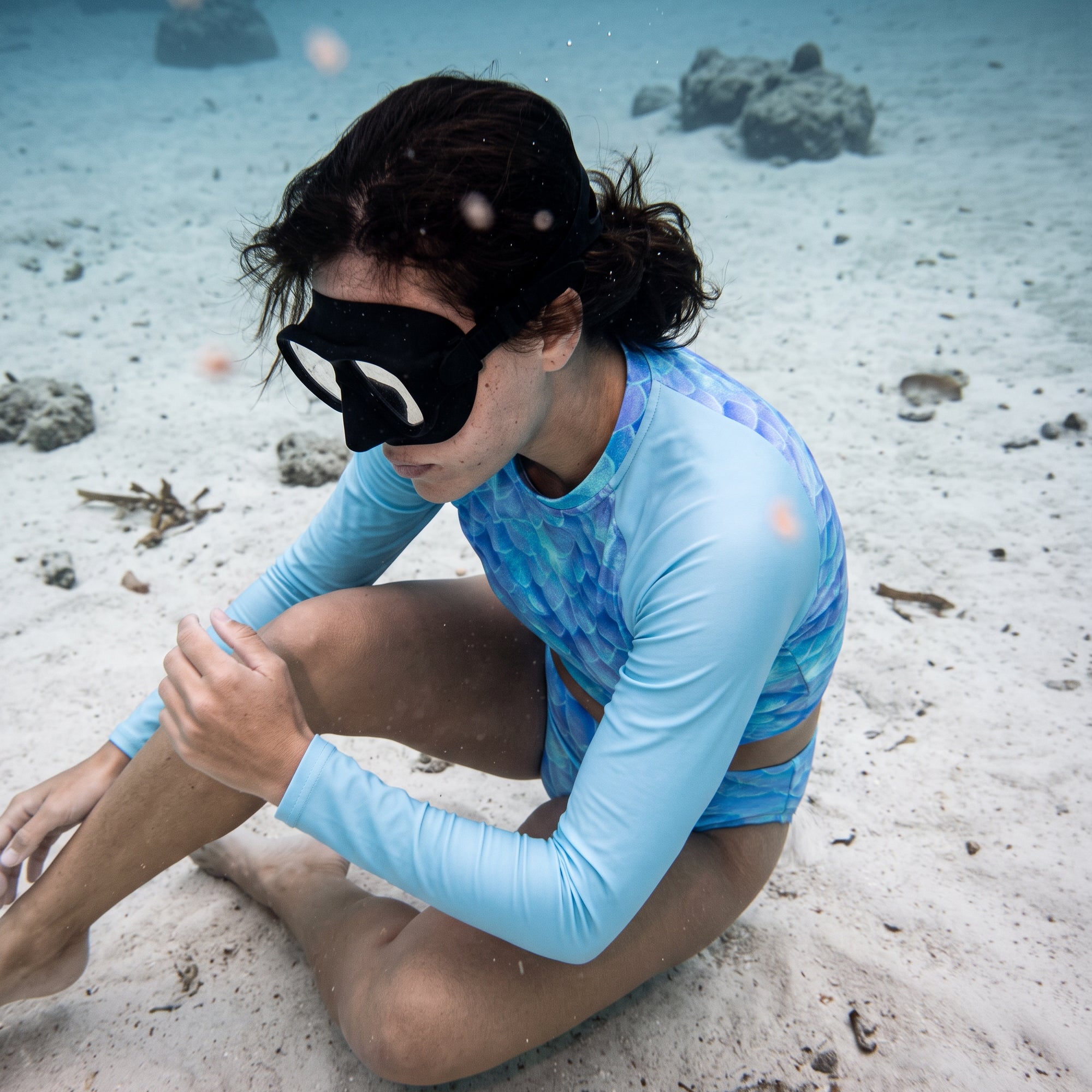 A woman underwater on a sandy seabed wearing a light blue long-sleeve swim shirt and matching blue patterned swim bottoms, with a black swim mask.