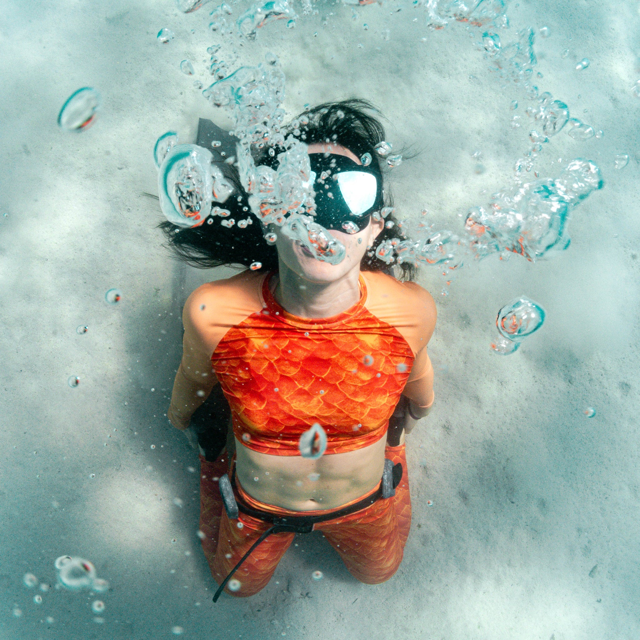 A person underwater wearing a bright orange-red fish-scale patterned bikini top and matching bottoms, with a black swim cap and goggles, surrounded by bubbles and clear water.