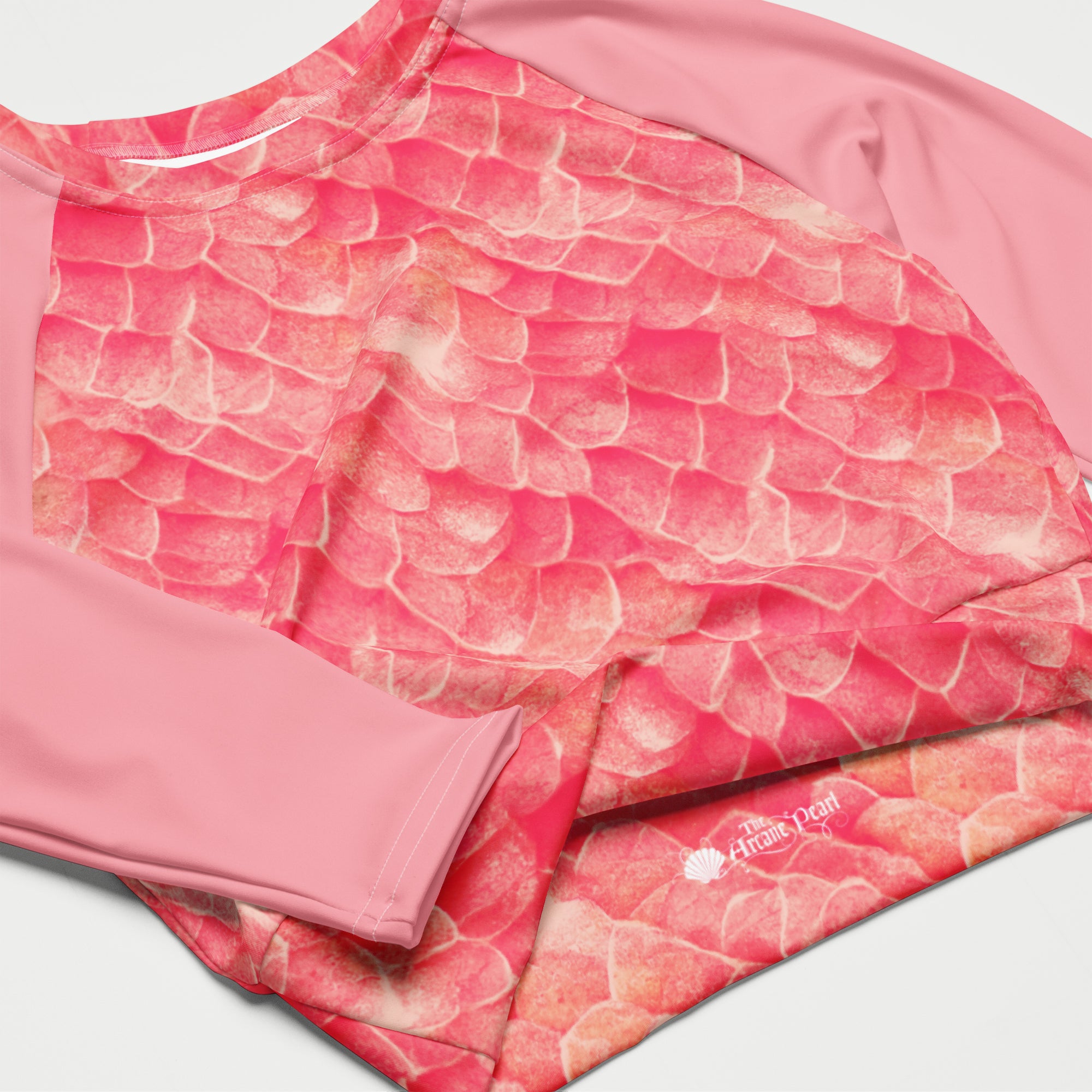 A close-up of a pink long-sleeve athletic top with a textured, scale-like pattern in shades of coral, pink, and peach, featuring solid pink sleeves and a small white logo near the hem.